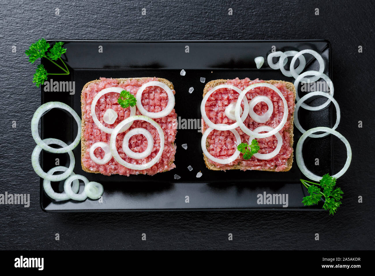 Deutsche essen mett Boden Schweinefleisch, Bun- und rohes Fleisch mit Zwiebeln und Petersilie auf schwarzem Hintergrund Stockfoto