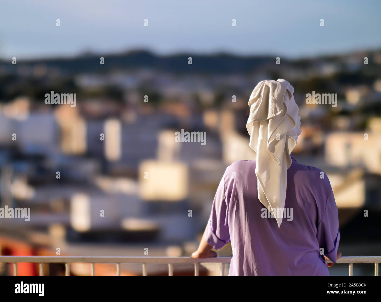 Eine Frau steht auf einem Balkon am Morgen mit einem weißen Handtuch auf dem Kopf vor dem Hintergrund der Stadt, Ansicht von hinten Stockfoto