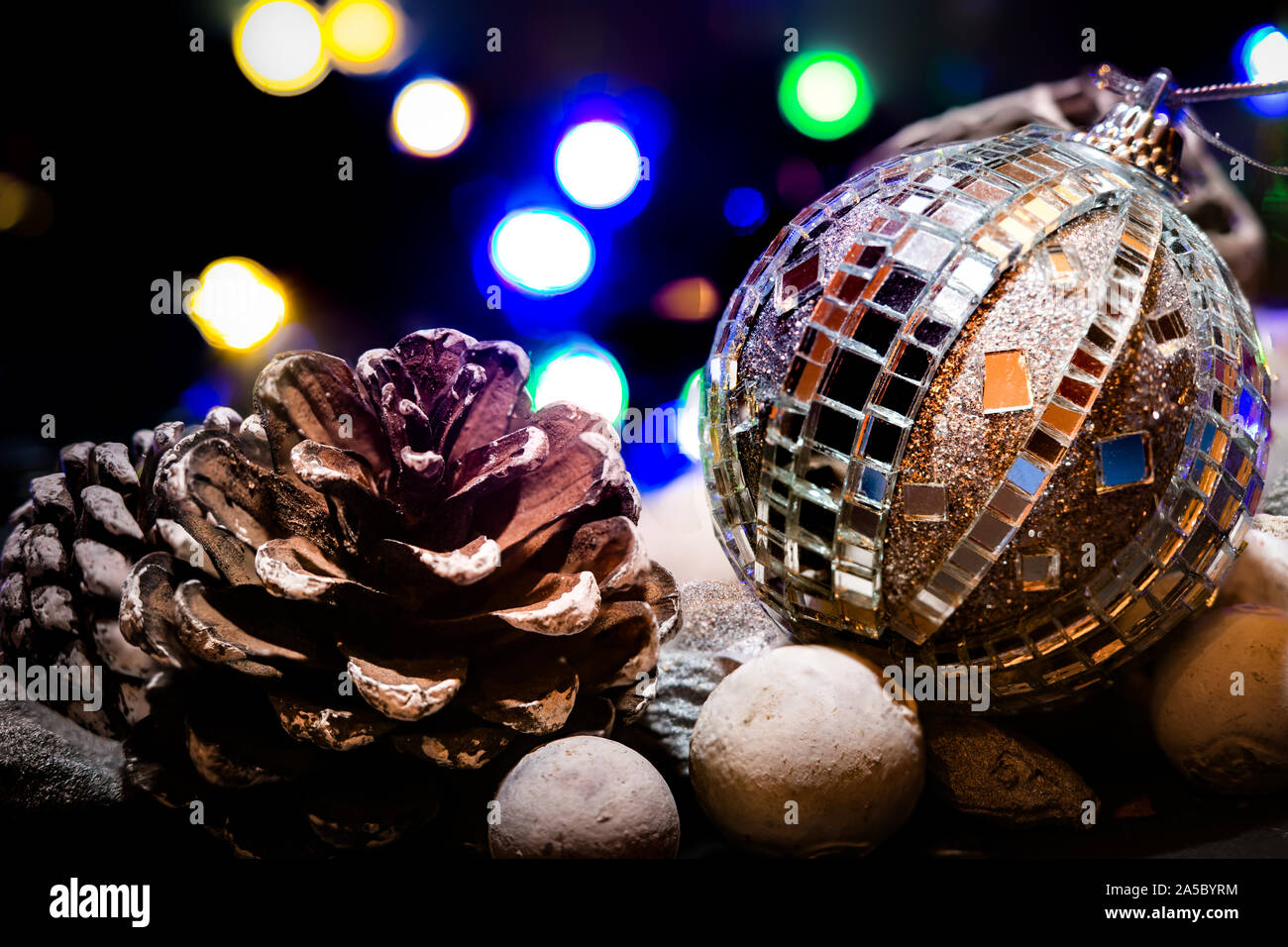 Weihnachtsschmuck Szene mit Lichterketten vor einem schwarzen Hintergrund. Eine silberne Christbaumkugel mit Spiegel sitzt neben zwei Tannen Zapfen auf einem Bett aus Kieselsteinen. Stockfoto