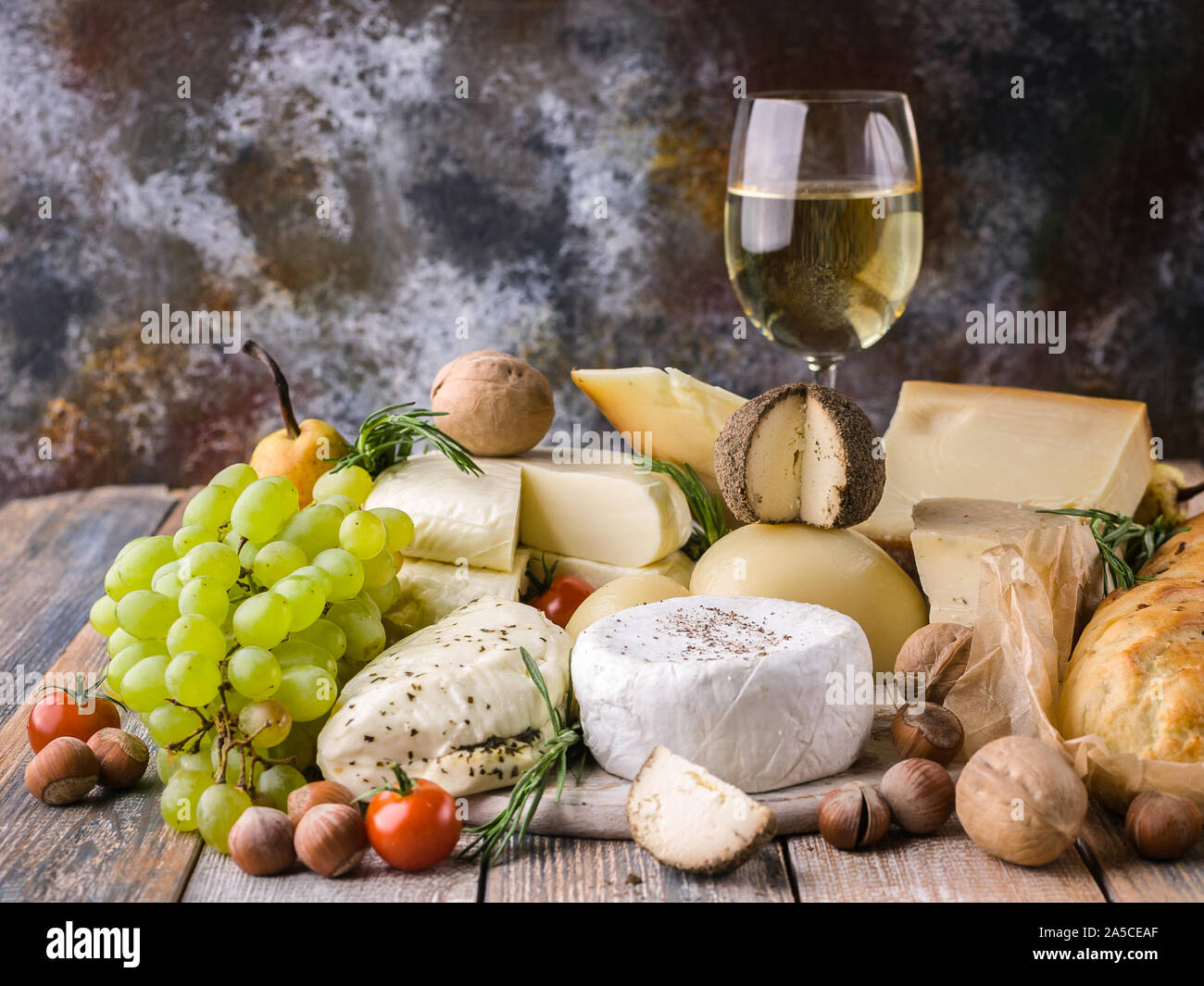 Essen still life: Verschiedene köstliche Käsesorten, Glas Weißwein, Nüssen, Obst, würzige Kräuter und Zwiebeln baguette auf einem urigen Hintergrund. Stockfoto