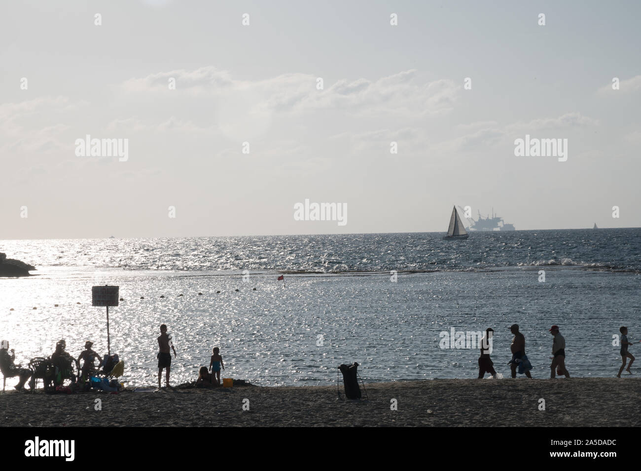 DOR STRAND, Israel/18 OKT 2019: Badegäste am Strand als Segelboot an der Vorderseite der ökologisch umstrittene offshore Bohrinsel Stockfoto