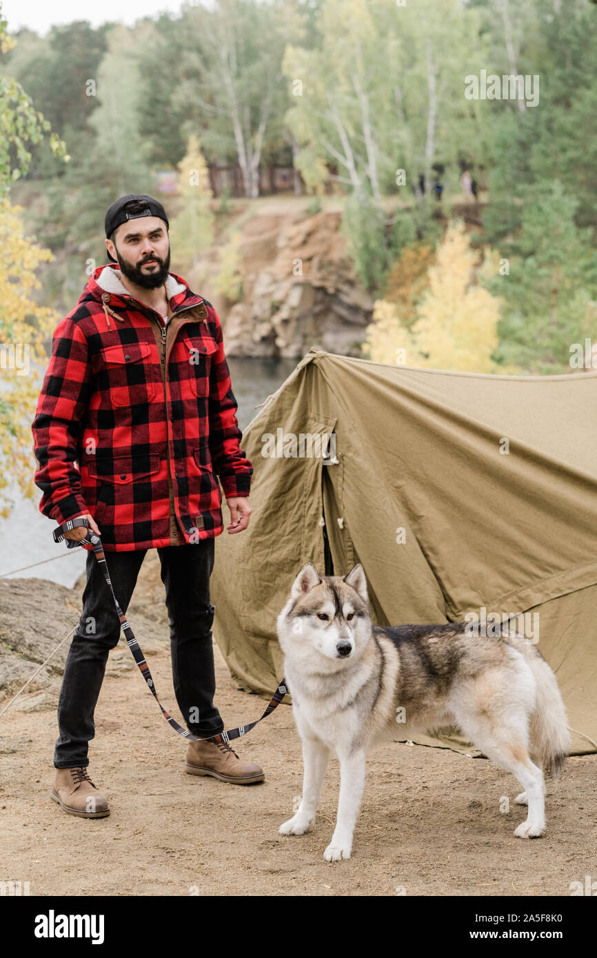 Junge Wanderer in casualwear Holding seinen süßen Husky PET-Leine steht die Hütte in ländlicher Umgebung Stockfoto