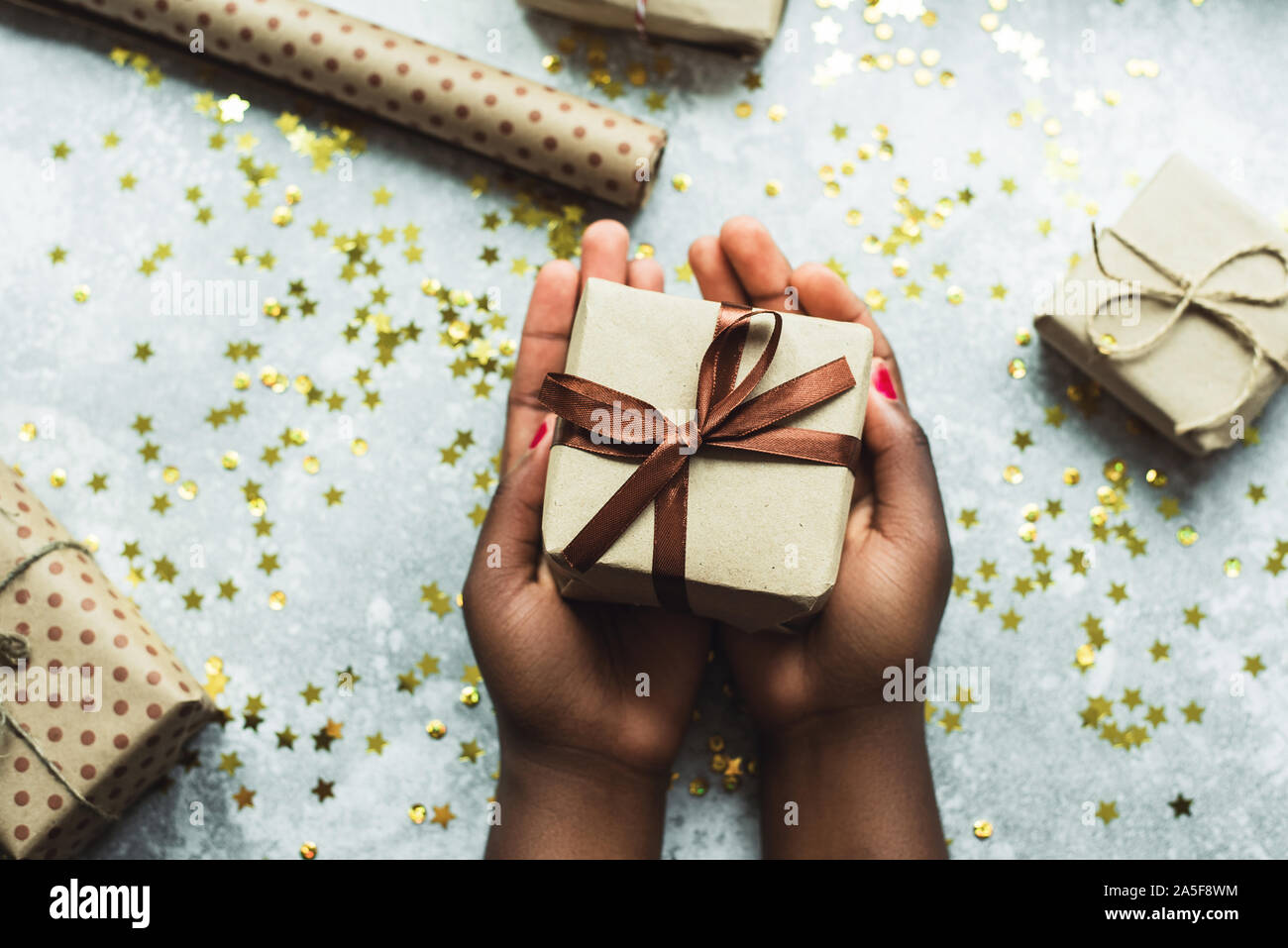 Eine Dunkelhautige Madchen Gibt Ein Geschenk Mit Ihren Eigenen Handen Gemacht Hande Halten Ein Handwerk Geschenk Mit Einem Braunen Band Auf Einem Grauen Hintergrund Mit Ein Paar Kleine Geschenke Flach Stockfotografie