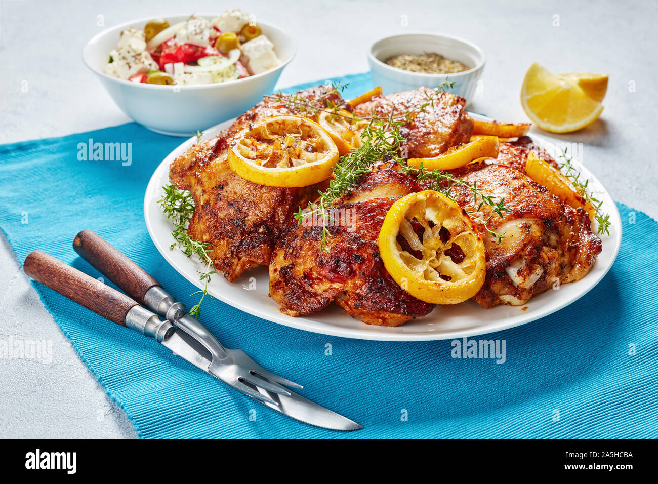 Knusprig gebratene saftige Hühnerkeule mit gerösteten Zitronenscheiben auf einer weißen Platte auf einer konkreten Tabelle mit Mediterraner Salat in eine Schüssel, Ansicht von oben Stockfoto
