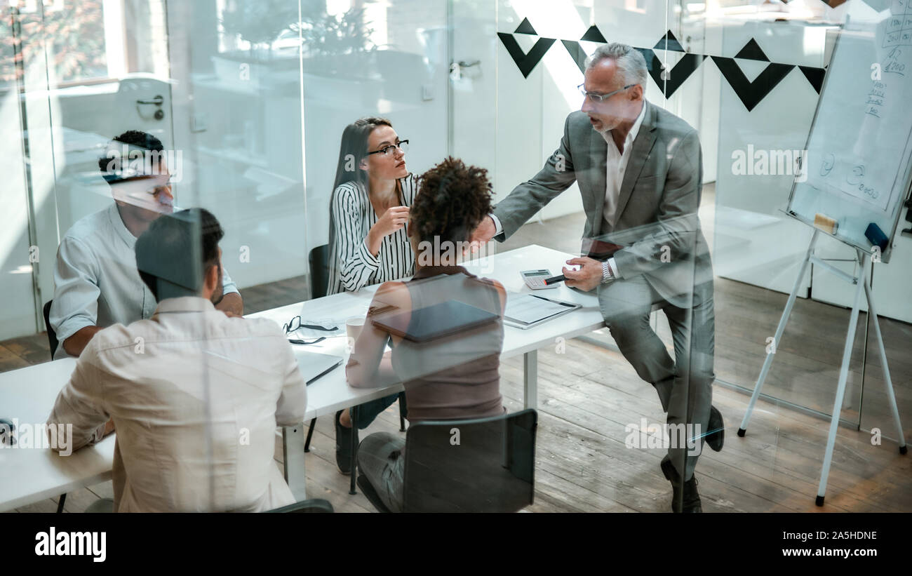 Geschäftsleute. Zuversichtlich reifer Mann in formalen Verschleiß erklärt etwas zu seinen Kollegen während der Sitzung auf dem Büro Tisch hinter der Glaswand in Stockfoto