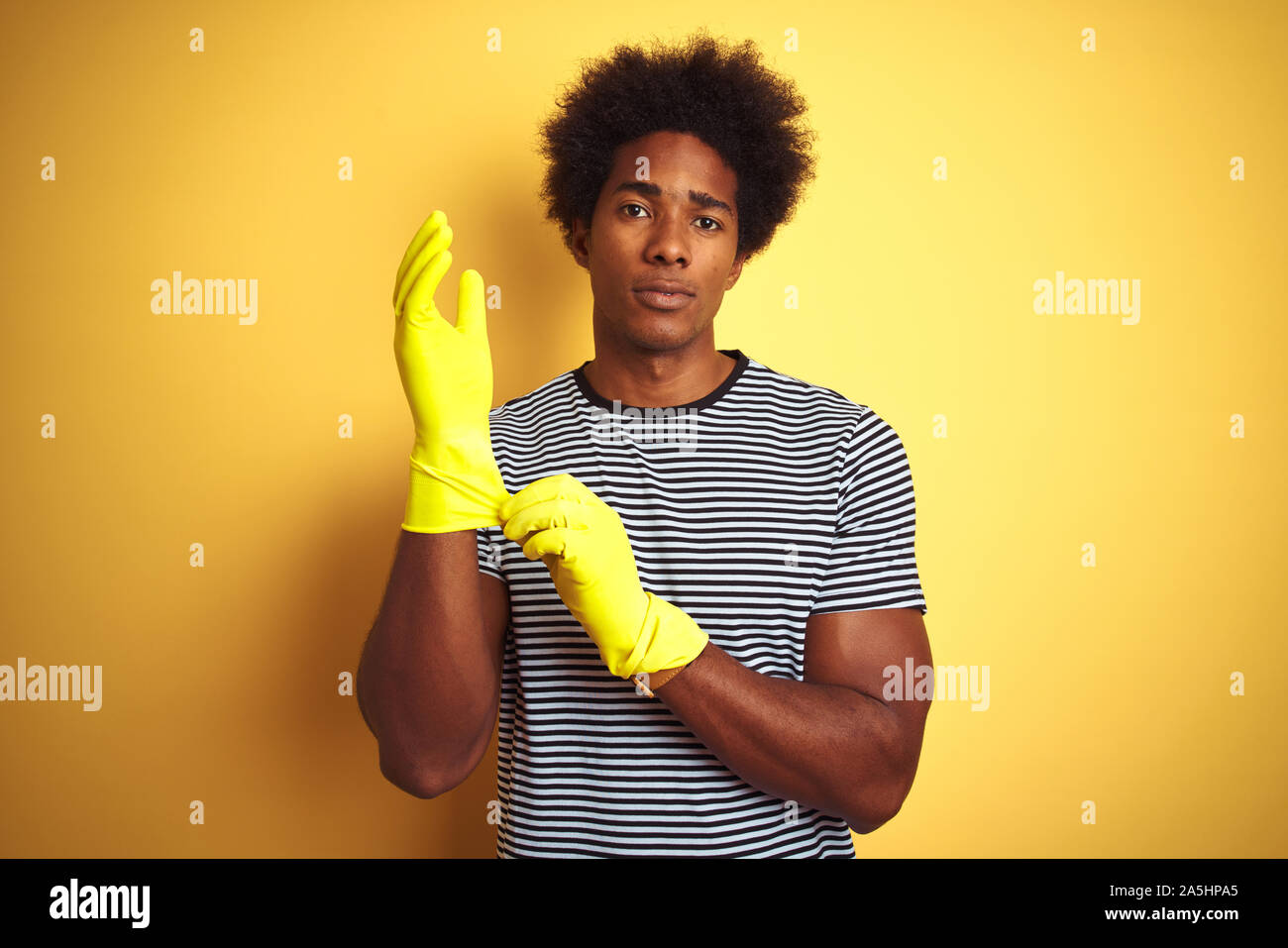 Junge afrikanische amerikanische Mann Reinigung mit Handschuhen über isolierte gelben Hintergrund stehend mit einem selbstbewussten Ausdruck auf Smart Face denken Ernst Stockfoto