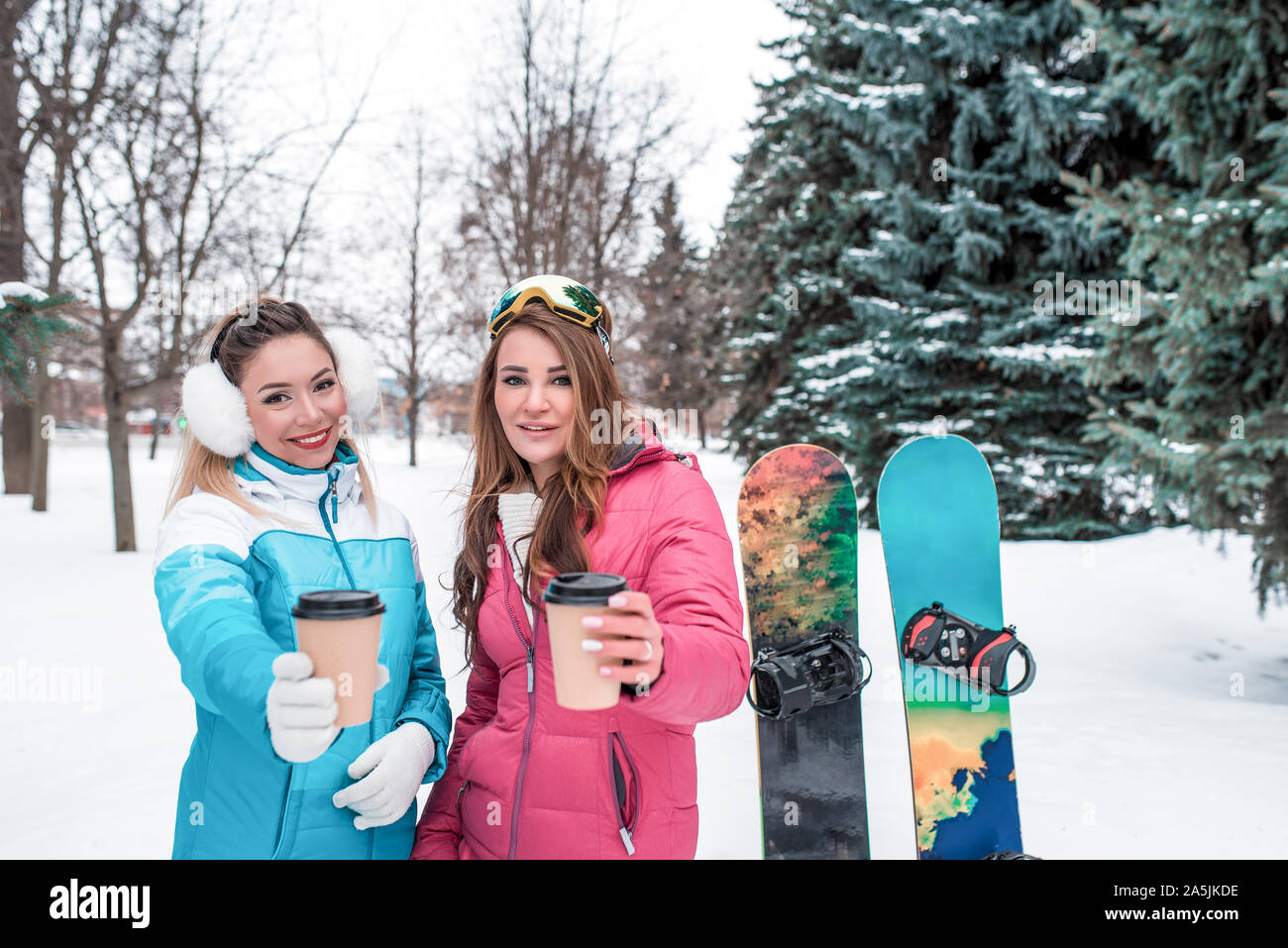 Zwei Mädchen Freundinnen Winter Park Resort, Tassen heißen Kaffee Tee, anbieten und mit Ihnen teilen. Emotionen Freude Komfort. Sport Overalls Stockfoto