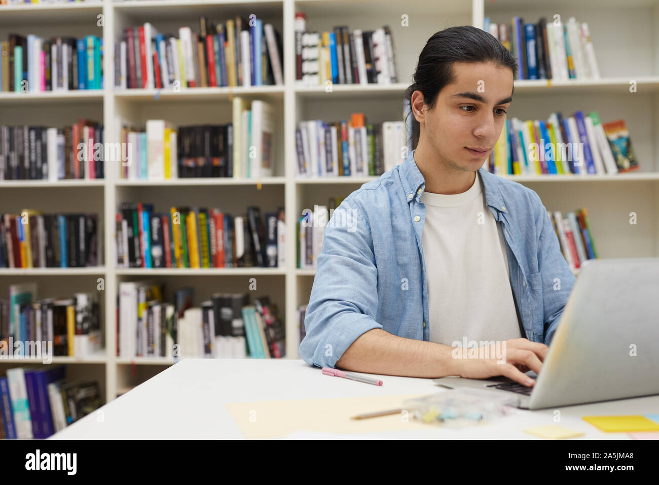 Portrait von jungen lateinamerikanischen Mann mit Laptop während des Studiums in der Hochschule Bibliothek sitzen gegen Bücherregale, Kopie Raum Stockfoto