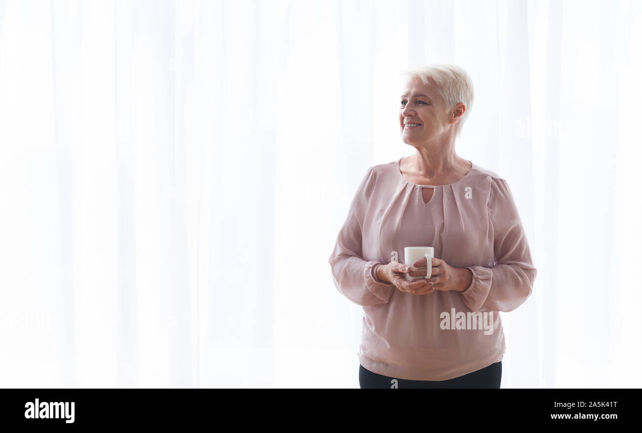 Nachdenklich alte Dame mit Becher bleiben neben Fenster Stockfoto