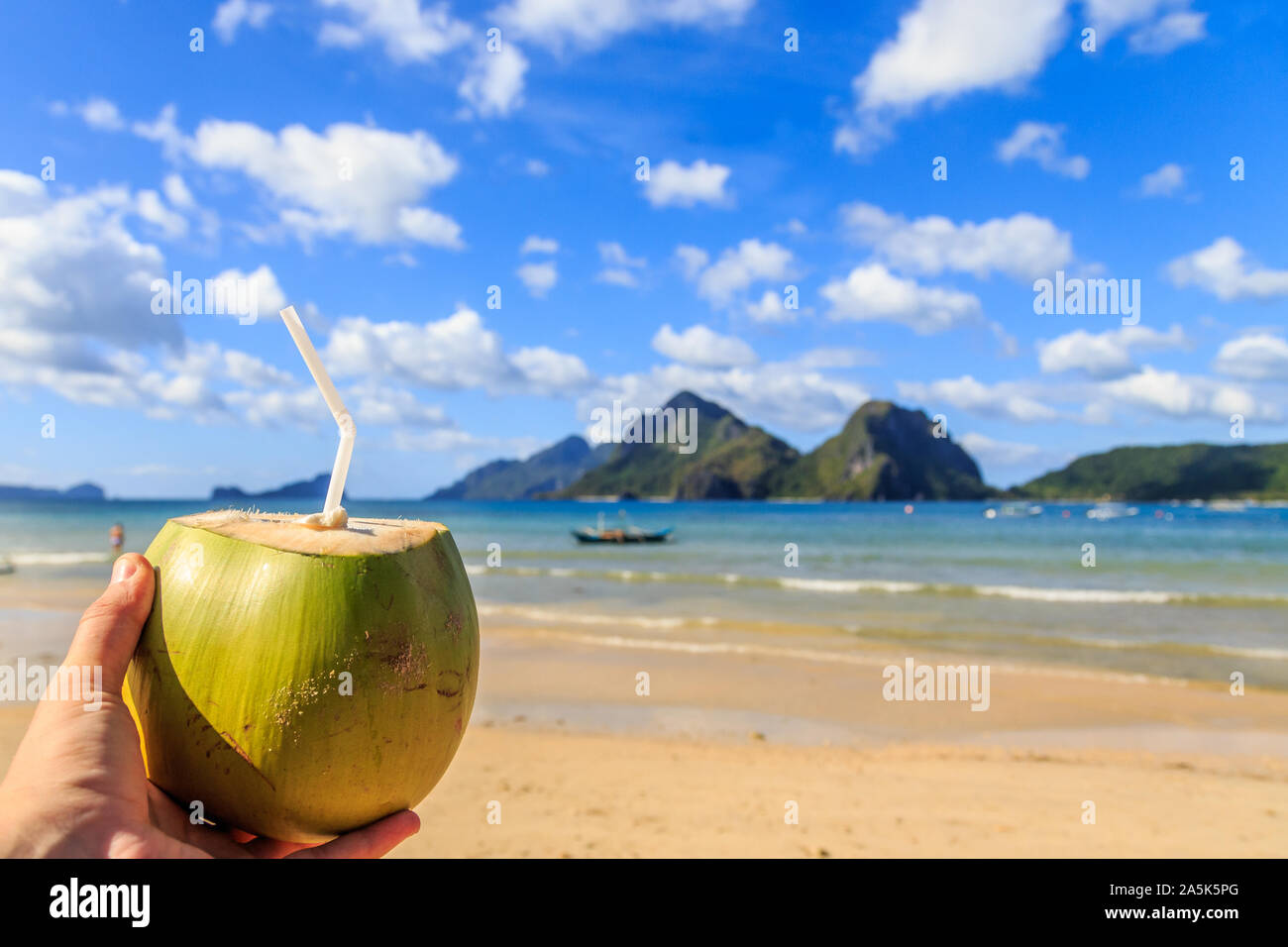 Hand mit einem Coconut coco loco Cocktails mit Stroh, tropische Inseln, Strand, Meer, blauer Himmel im Hintergrund, El Nido, Palawan, Philippinen Stockfoto