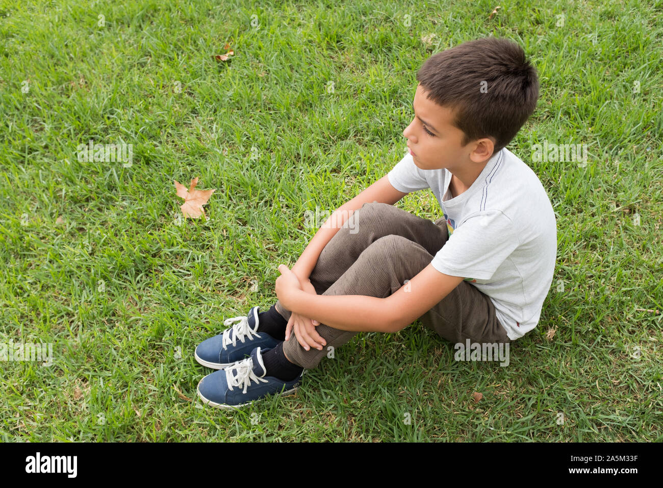 Seitenansicht von Konzentriertem braune Haare Junge mit verschränkten Armen allein sitzen auf dem Gras. Trauriges Kind weg schauen. Herbst Blätter im Hintergrund. Stockfoto