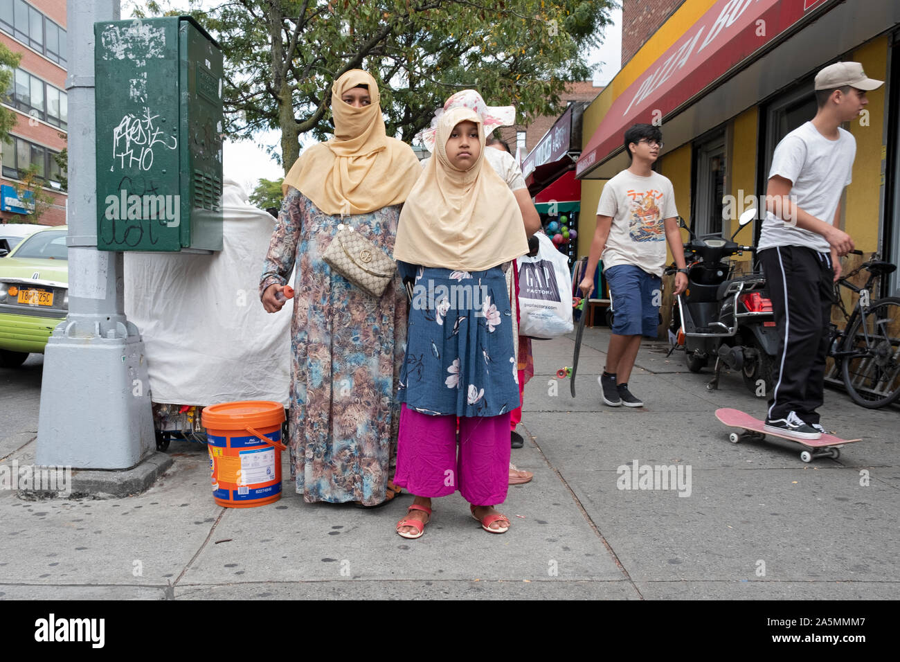 Eine Straßenszene in Jackson Heights mit 2 Frauen in traditionellen muslimischen Kleid und ein junger Mann auf einem Skateboard. In Queens, New York City. Stockfoto