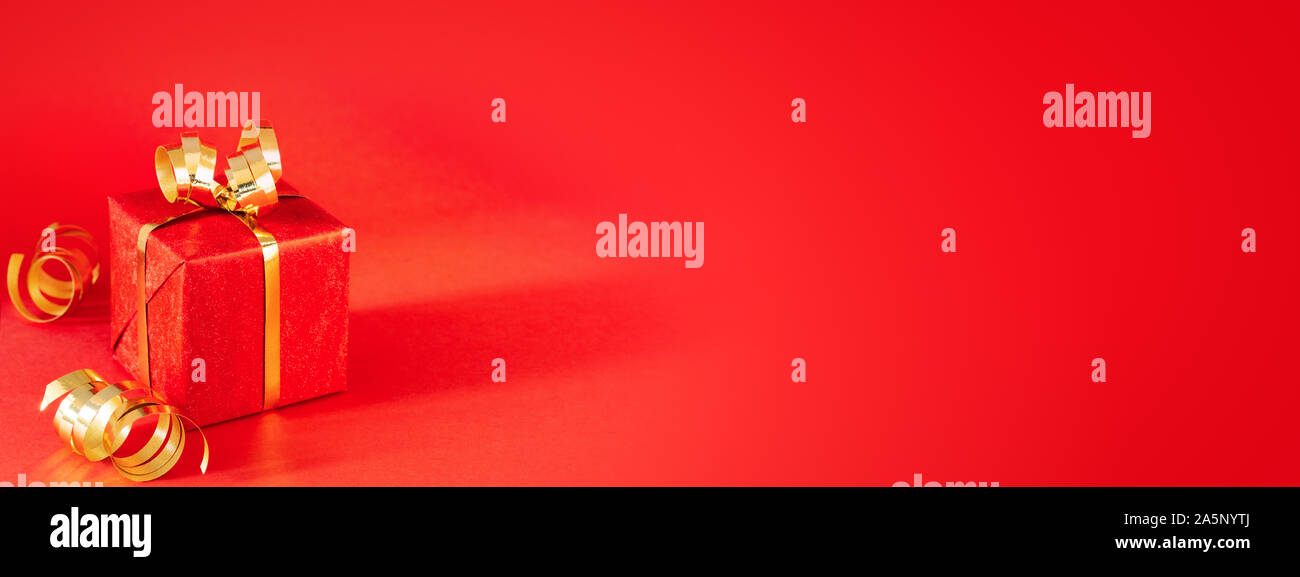 Roter Geschenkbox mit goldenen Bogen und Streamer auf rotem Hintergrund. Minimale Weihnachten und Geburtstag Banner. Schuß im Winkel. Stockfoto