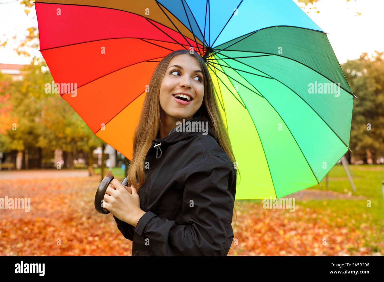 Fröhliche junge Frau mit bunten Regenschirm wieder ein Lächeln. Mädchen im Park mit Regenjacke und Schirm im Herbst. Stockfoto