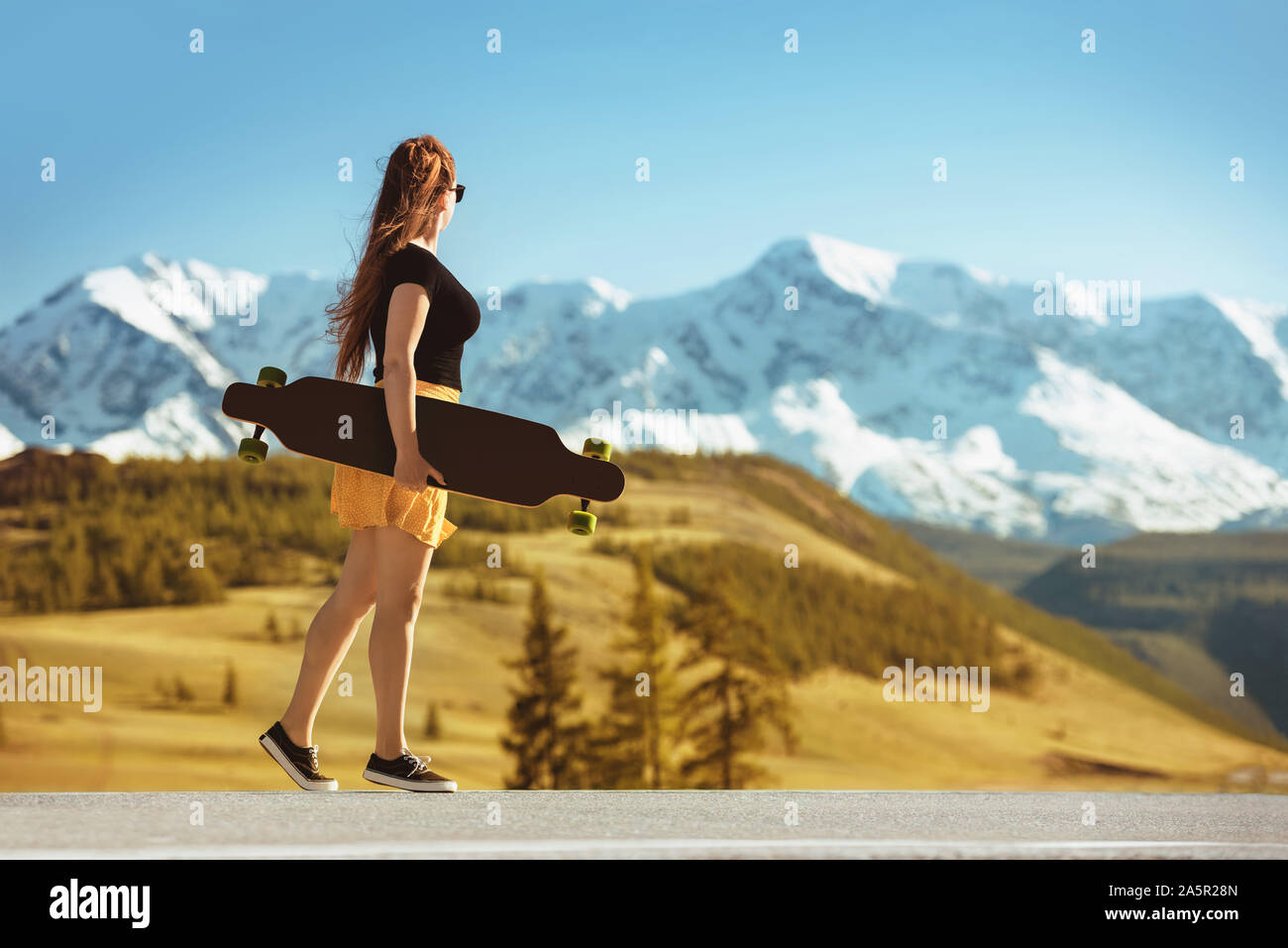 Junge Mädchen steht mit Longboard auf gerader Straße gegen Berge Stockfoto