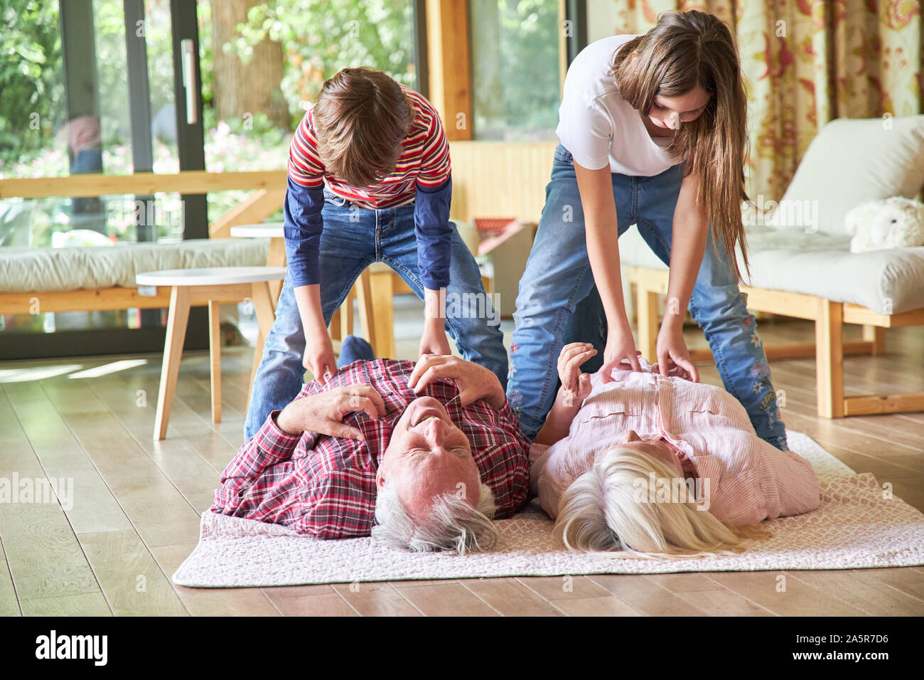 Zwei Geschwister Kinder kitzeln Oma und Opa auf dem Fußboden im Wohnzimmer Stockfoto