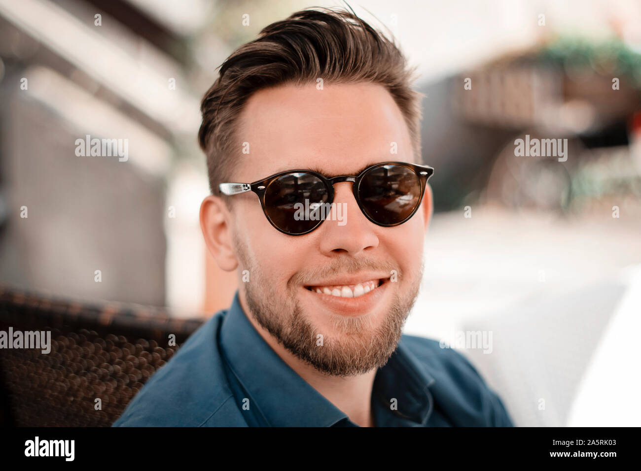 Porträt der jungen gutaussehenden Mann in Teal Blue Shirt mit Sonnenbrille und trendy Frisur. Mann lächelt und schaut in die Kamera draußen in einem Café. Stockfoto