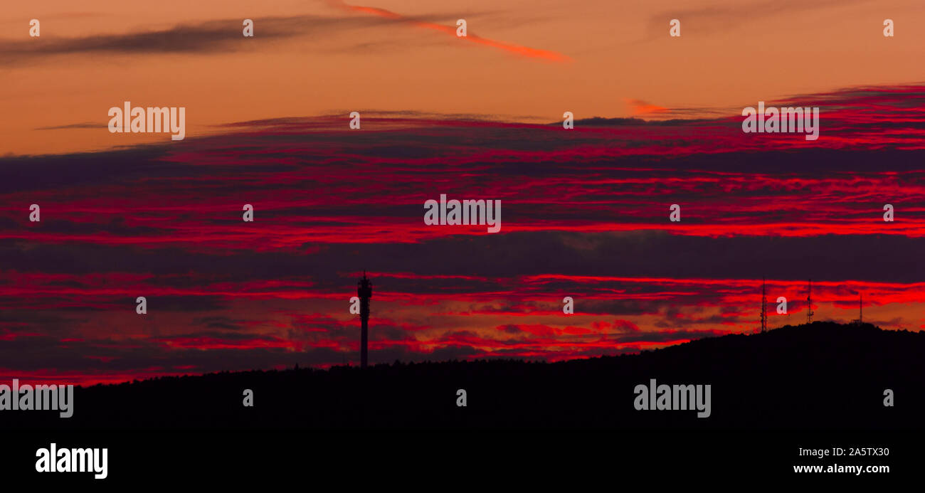 Sonnenuntergang hinter einem Hügel. Große Wolken in Rot und Orange. Schwarze Silhouetten von Funktürmen auf dem Hügel. Stockfoto