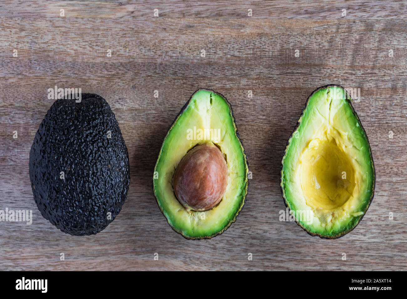 Eine ganze und eine halbierte Avocado mit der Grube in der Mitte Früchte. Die avocados sind auf einer hölzernen Hintergrund. Die Kamera wird direkt von oben, Fl Stockfoto