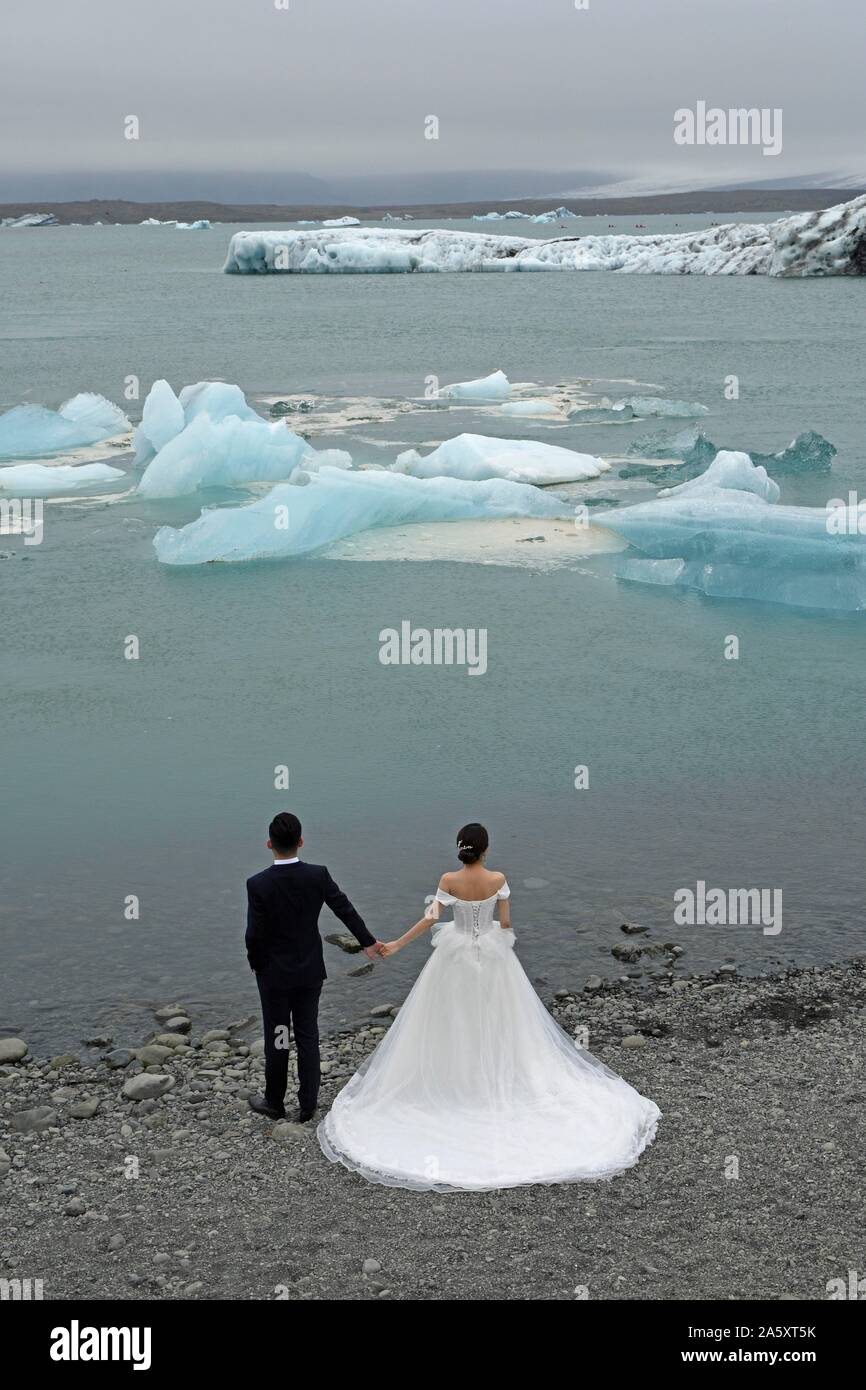 Braut in einem klassischen weißen Brautkleid und Bräutigam im dunklen Anzug mit Blick auf schwimmenden Eisbrocken und die Gletscher am Ufer des Stockfoto