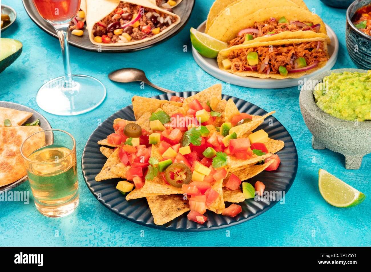 Mexikanisches Essen mit Nachos vorne und Taco shells mit gezogenen Schweinefleisch, Burritos, Guacamole und andere Gerichte, mit Tequila und Kalk auf einem blauen Hintergrund Stockfoto