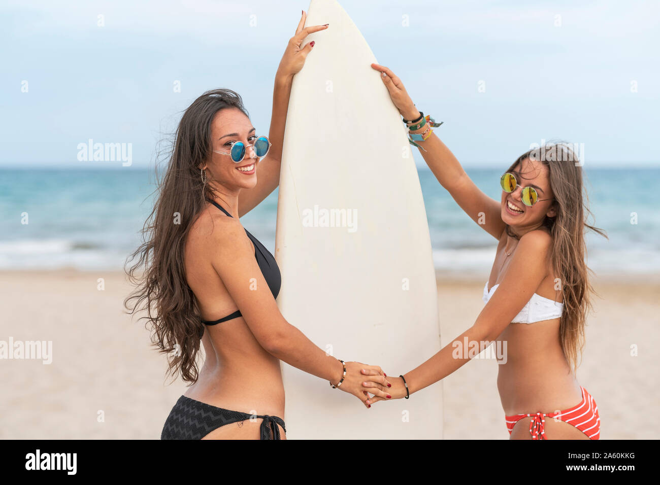 Zwei glückliche weibliche Freunde hand in hand am Strand mit Surfbrett Stockfoto