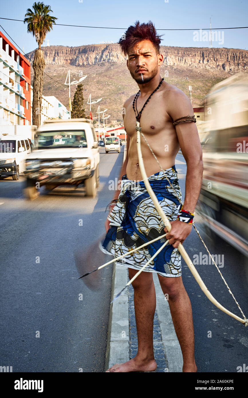 Tribal Mann mit seinem traditionellen Bogen und Pfeile in der Mitte der Stadt mit Autos vorbei, Lubango, Angola Stockfoto