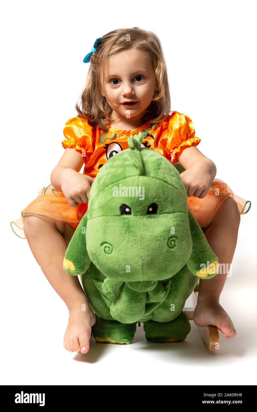 Ein kleines Mädchen in einem orangefarbenen Kleid vor einem weißen Hintergrund sitzt auf einem grünen rockigen Dinosaurier gekleidet Stockfoto