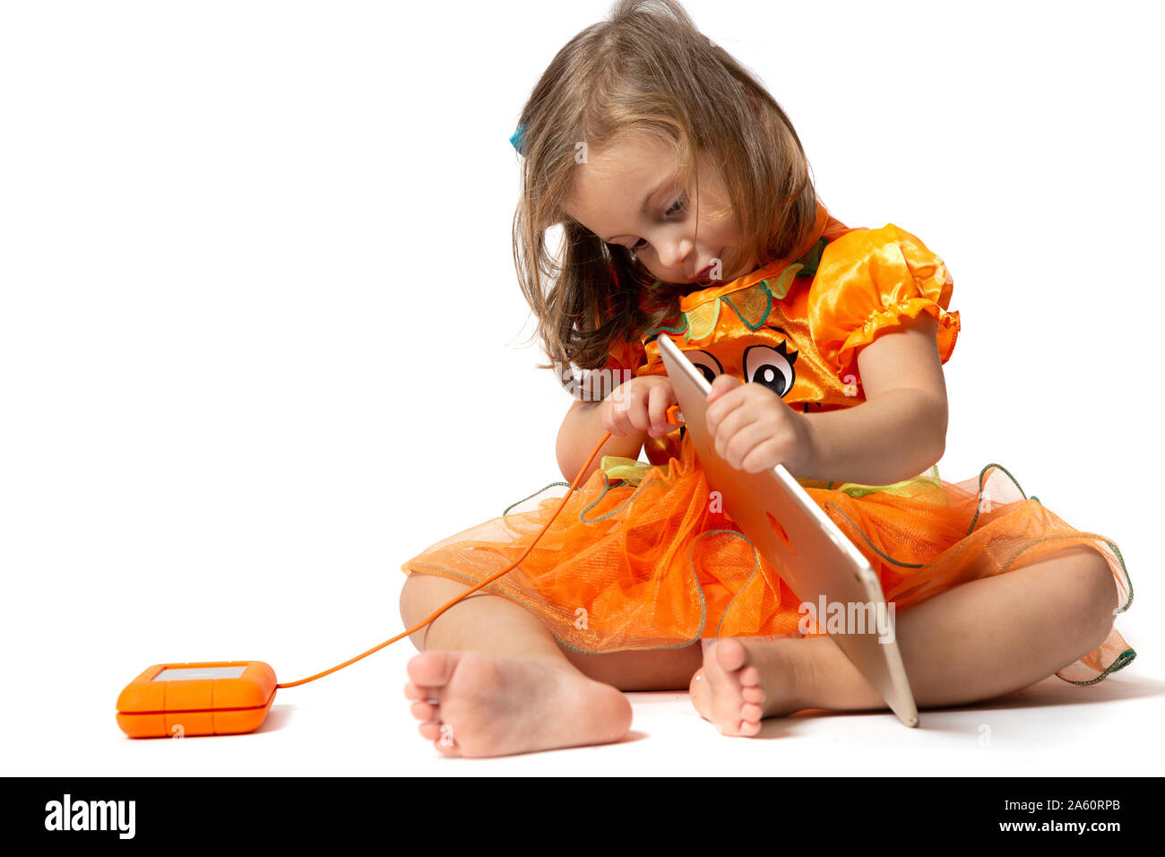 Ein kleines Mädchen in einem orangefarbenen Kleid, die versuchen, eine orange Festplatte auf eine Tablette zu verbinden gekleidet Stockfoto