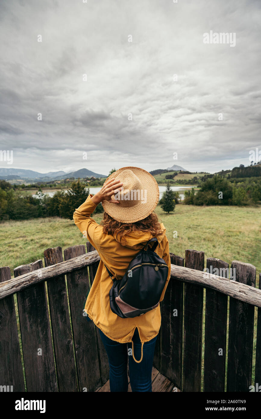 Junge Frau tragen gelbe Jacke und Rucksack mit einer Hand mit einem Hut in den Kopf schauen, um den See Landschaft auf einem Holz Balkon Stockfoto