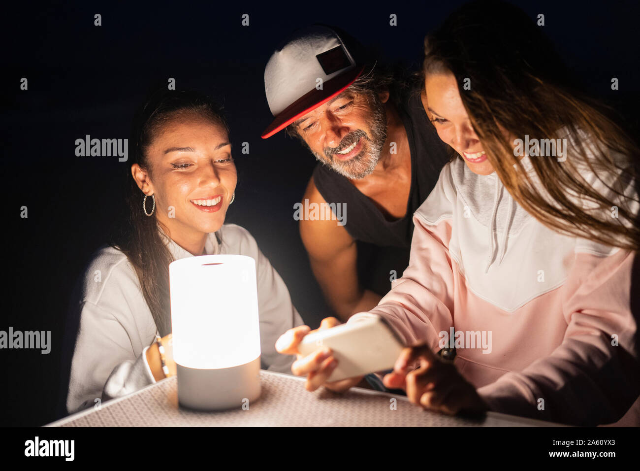 Zwei junge Frauen und reife Männer mit Smartphone und LED-Licht auf Camping Tisch bei Nacht Stockfoto