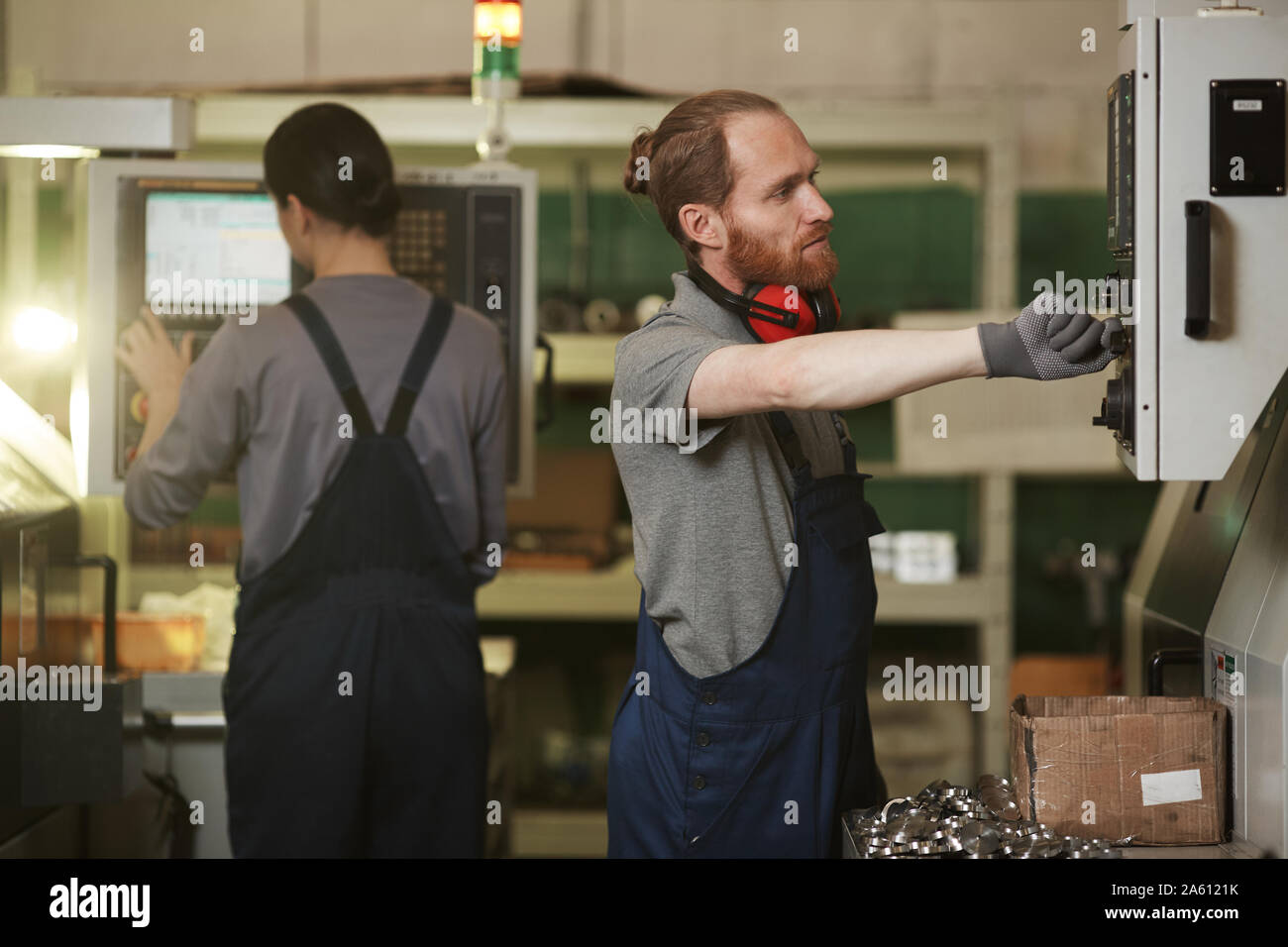 Junge bärtige Mann in Overalls stehen in der Nähe der Maschine und Kontrolle der Arbeit des es mit Frau im Hintergrund arbeiten Stockfoto