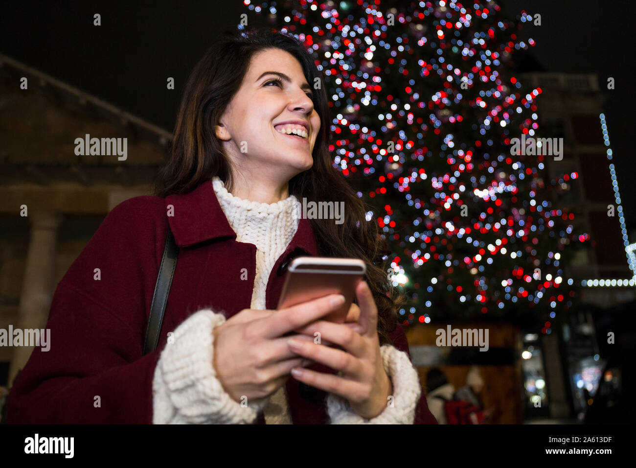 Porträt der glückliche junge Frau mit Smartphone vor der beleuchteten Weihnachtsbaum im Freien Stockfoto