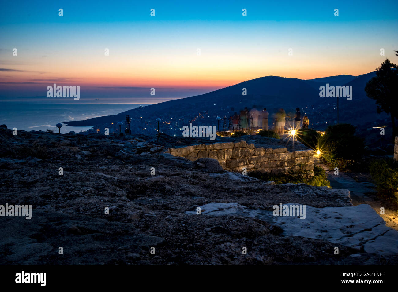 Blaue Stunde frühe Nacht Landschaft von Lekuresi Schloss, Saranda, Albanien mit Adria Küste, Clear Spring Sky mit Haze, Langzeitbelichtung Bewegung verwischt unkenntlich Menschen auf der Aussichtsplattform Stockfoto