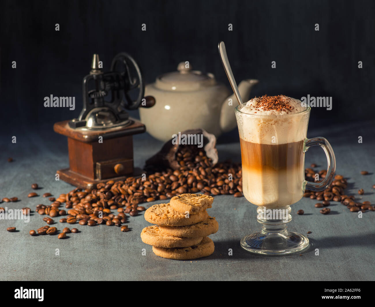 Hot Latte Macchiato in einem hohen Glas mit Schokolade Streusel auf der Oberseite. Im Hintergrund gibt es Kaffeebohnen verschüttet und eine Kaffeemühle. Stockfoto