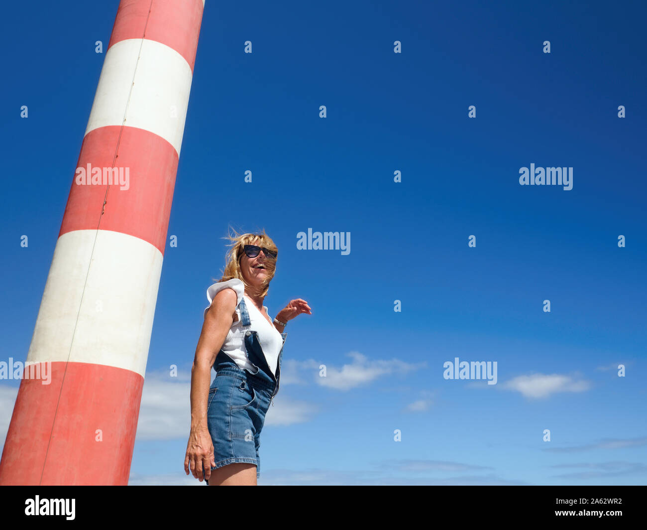 Eine junge Frau steht neben einem Teil einer rot-weiss gestreifte Leuchtturm, dahinter blauer Himmel und ein paar sehr kleine weiße Wolken. Sie trägt Hosen und Stockfoto