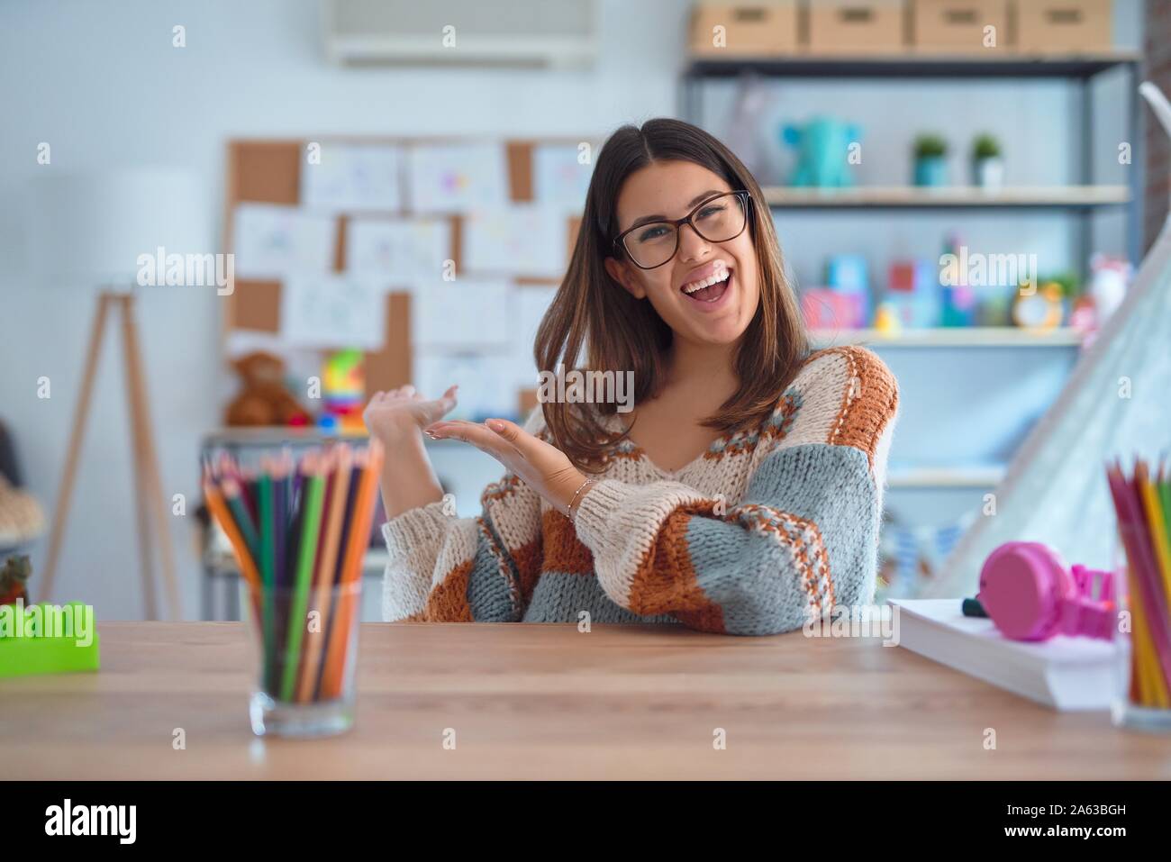 Junge schöne Lehrerin Frau mit Pullover und Gläsern sitzen am Schreibtisch im Kindergarten Einladenden natürlichen lächelnd mit offenen Hand eingeben Stockfoto