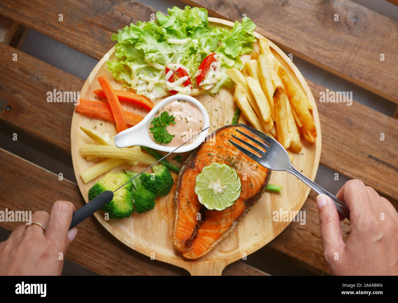 Lachs Steak serviert auf Holzplatte, Beilagen, gekochtes Gemüse, Pommes frites, Salat, Sauce und einem Stück Zitrone auf der Oberseite Stockfoto