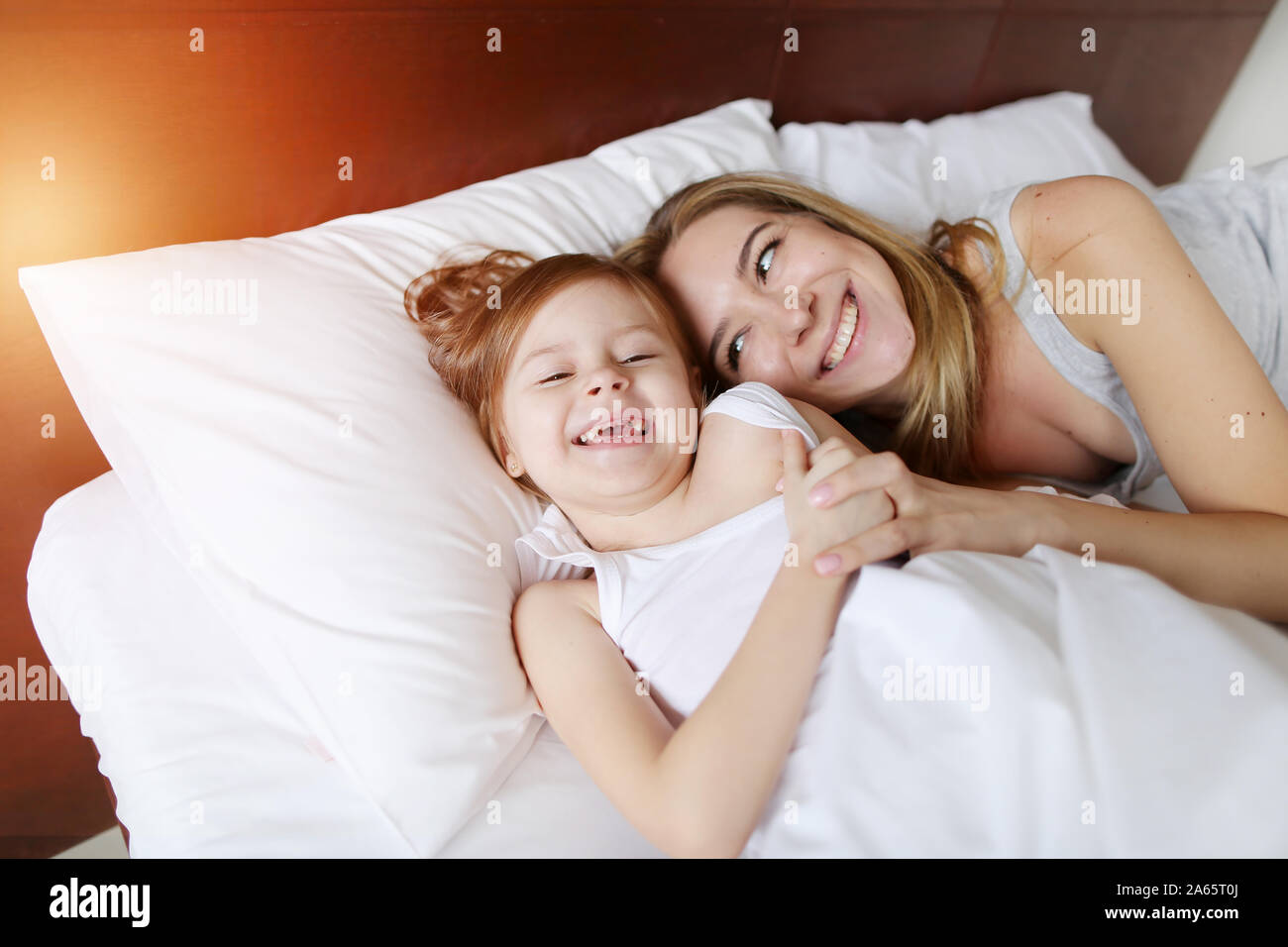 Schönheit Mutter umarmt kleine Tochter am weißen Bett mit Sonnenschein Stockfoto
