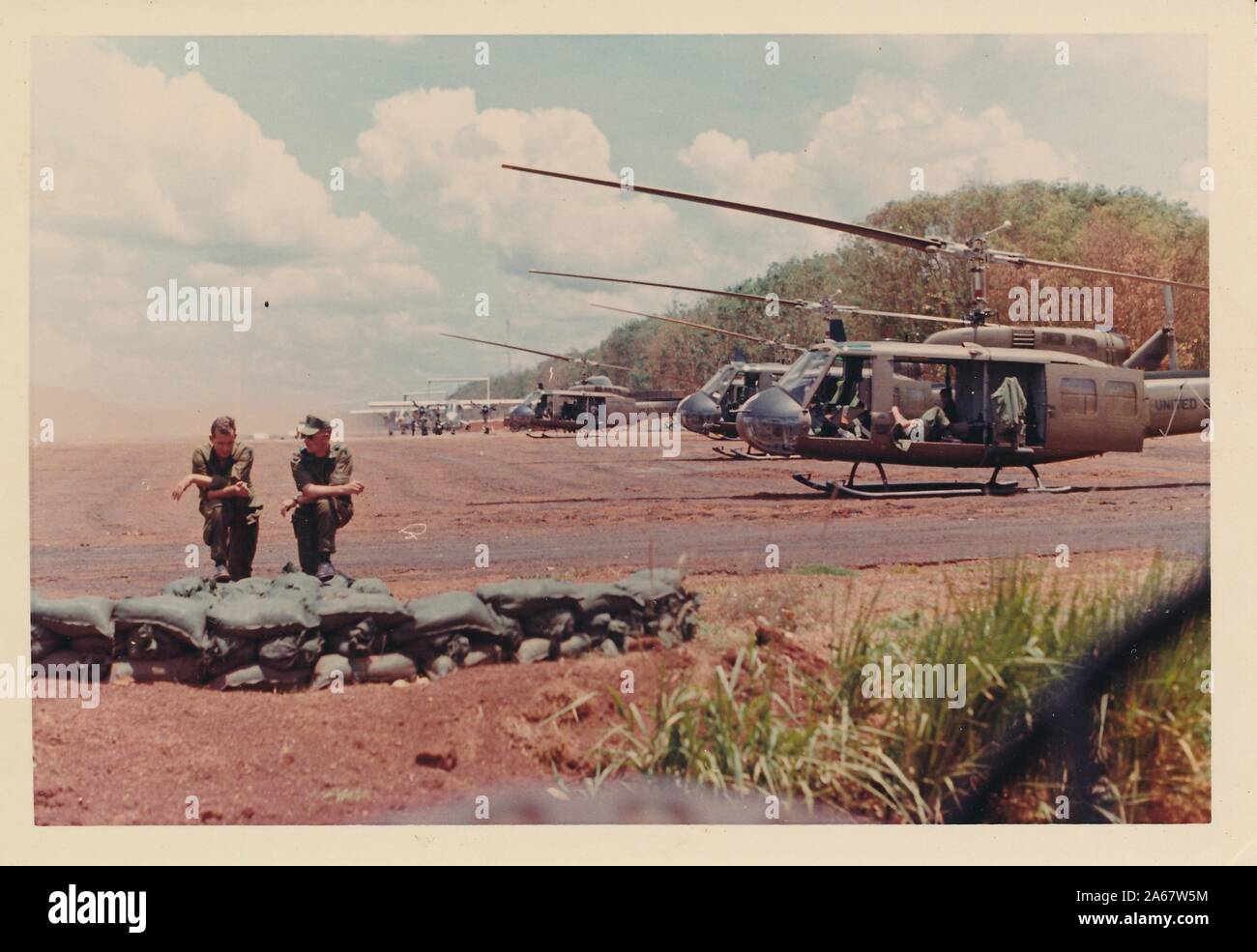 Zwei amerikanische Soldaten sprechen und ruhen in der Nähe eines Foxholes mit Sandsäcken, mit einer Reihe von Hubschraubern vom Typ Bell Uh-1 Iroquois (Huey) warten im Hintergrund, in Vietnam während des Vietnamkriegs 1975. () Stockfoto