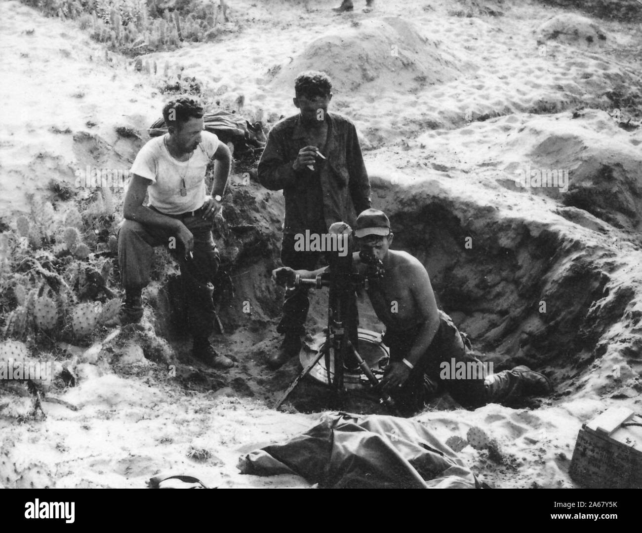 Ein hochwinkeliger Schuss von zwei amerikanischen Soldaten, die draußen in einem sandigen Dugout stehen und dabei zusehen, wie ein Kollege auf dem Boden kniet, um durch die Sehenswürdigkeiten der Stativpistole oder des Teleskops, Vietnam, 1965 zu schauen. () Stockfoto