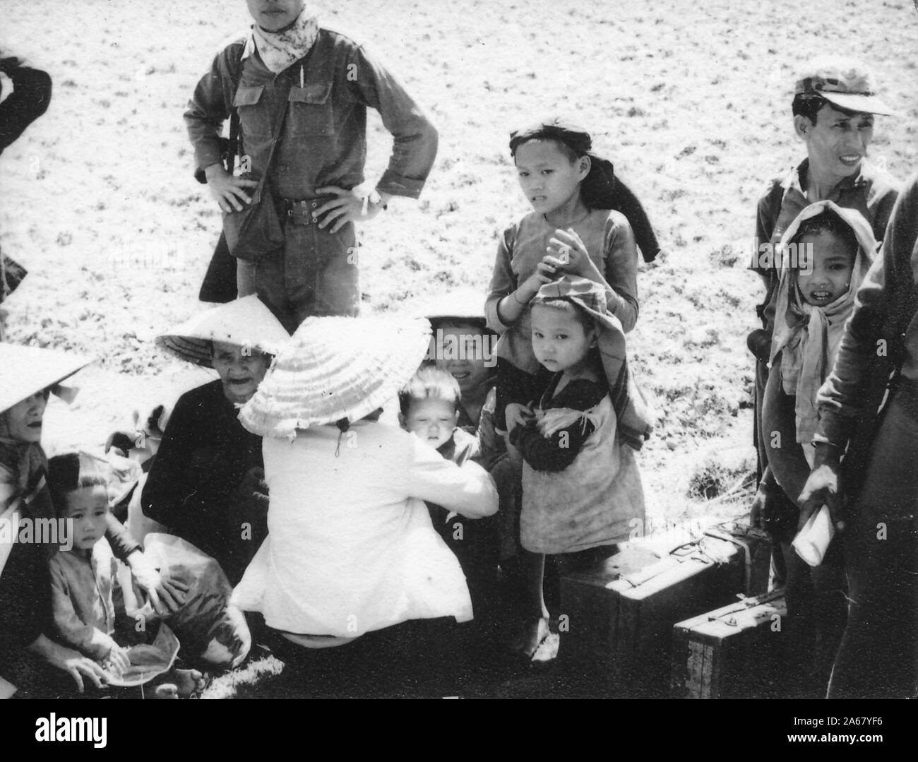 Hochwinkeliger Schuss einer kleinen Gruppe vietnamesischer Frauen und Kinder, draußen sitzend und stehend an einem sonnigen Tag, flankiert von Soldaten, Vietnam, 1965. () Stockfoto