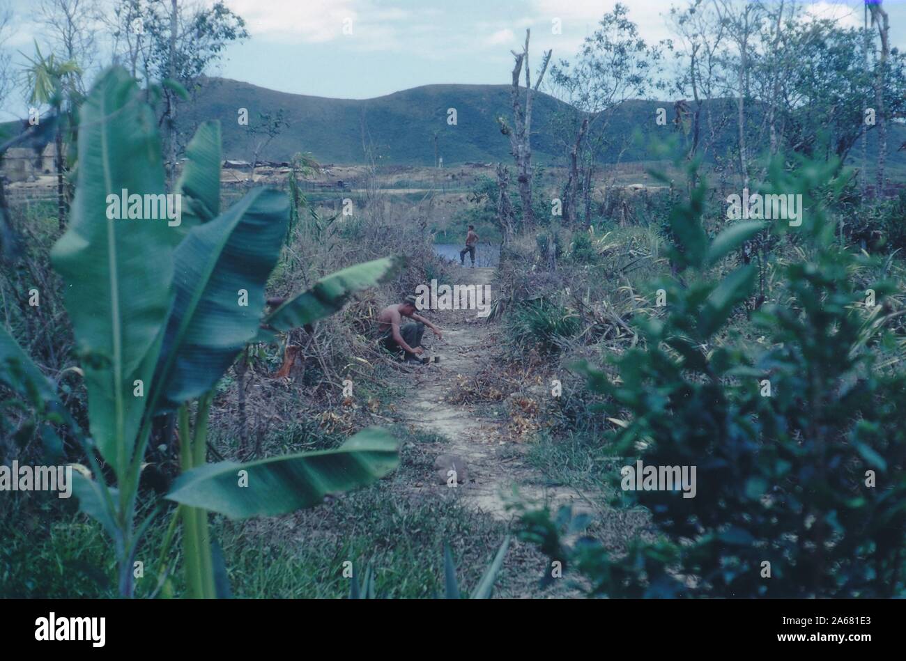 Ein langer Schuss von einem schmutzigen Pfad, der zu einem Gewässer in einem ländlichen Gebiet führt, wobei ein Soldat auf dem mittleren Pfad sitzt und ein anderer in der Nähe des Wasserrandes, Vietnam, 1965, steht. () Stockfoto