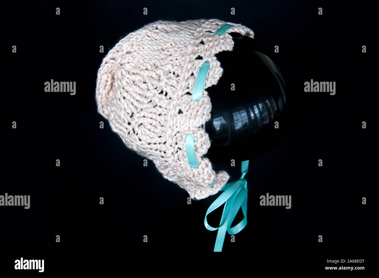 Ecru knit baby Haube auf einem schwarzen Hintergrund. Spitze Zubehör für Säuglinge mit einem Aqua farbiges Satinband dieses Handwerk gestrickt. Stockfoto