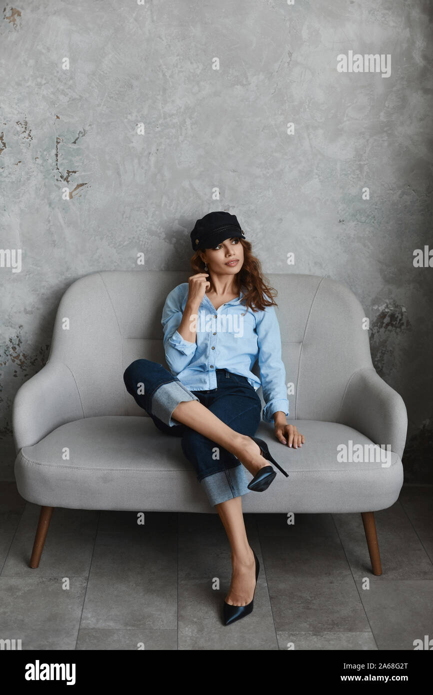 Hohe altmodische Modell Mädchen mit High Heel Schuhe, Hut, blaues Hemd und Roll-up Jeans. Schöne junge Frau in modischen Outfit sitzt auf der Couch und Stockfoto