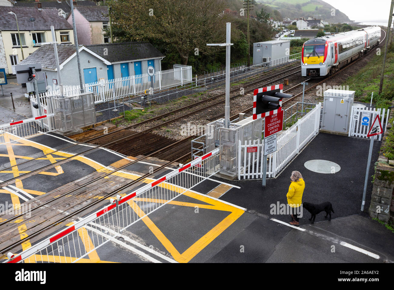 Frau in gelbem Mantel wartet an einem Bahnübergang in Ferryside, Wales, als ein Zug sich nähert Stockfoto