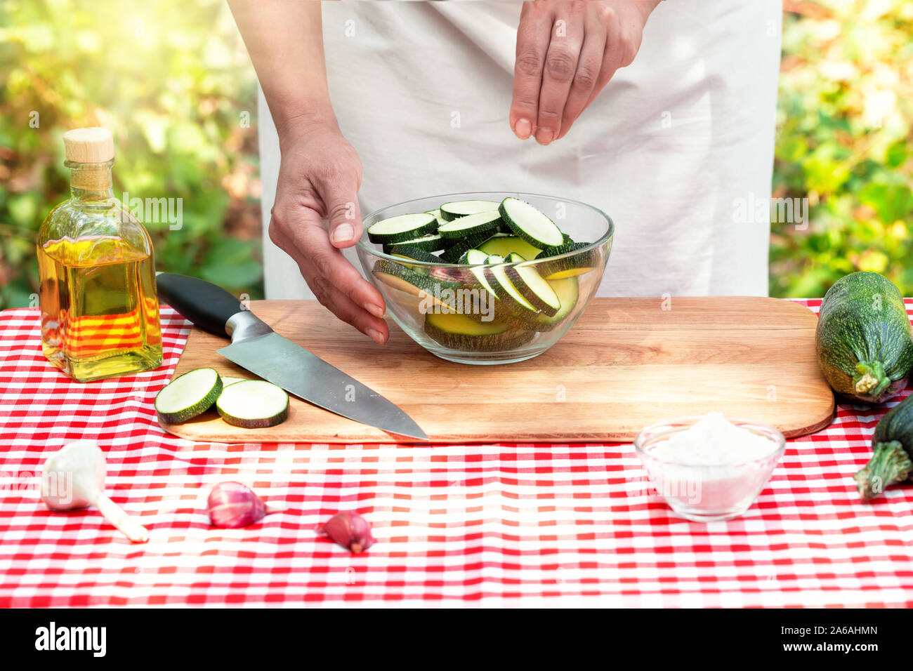 Weibliche Kochen Salz geschnittene Zucchini in einem transparenten Schüssel. Karierte Tischdecke auf dem Tisch, natürlichen, grünen Hintergrund. Das Konzept des Kochens natürliche Eg Stockfoto