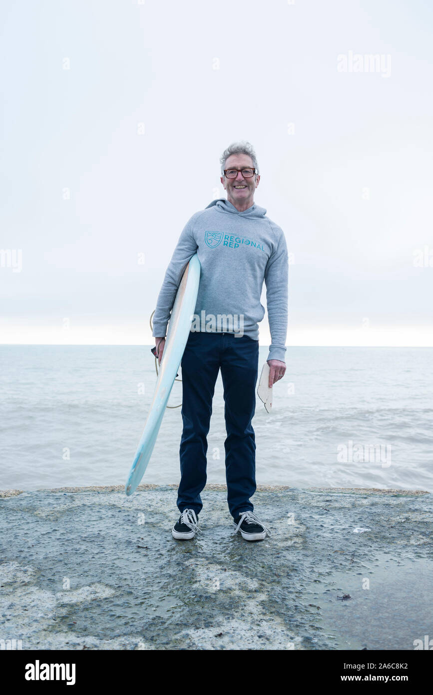 Ein reifer Mann mit dem grauen Haar steht am Wasser mit einem Surfbrett Stockfoto