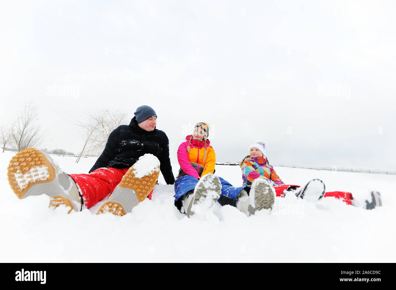 Eine junge Familie - Mutter, Vater und Tochter in hellen Skianzuege sitzen zusammen im Schnee Lachen und Lächeln Stockfoto