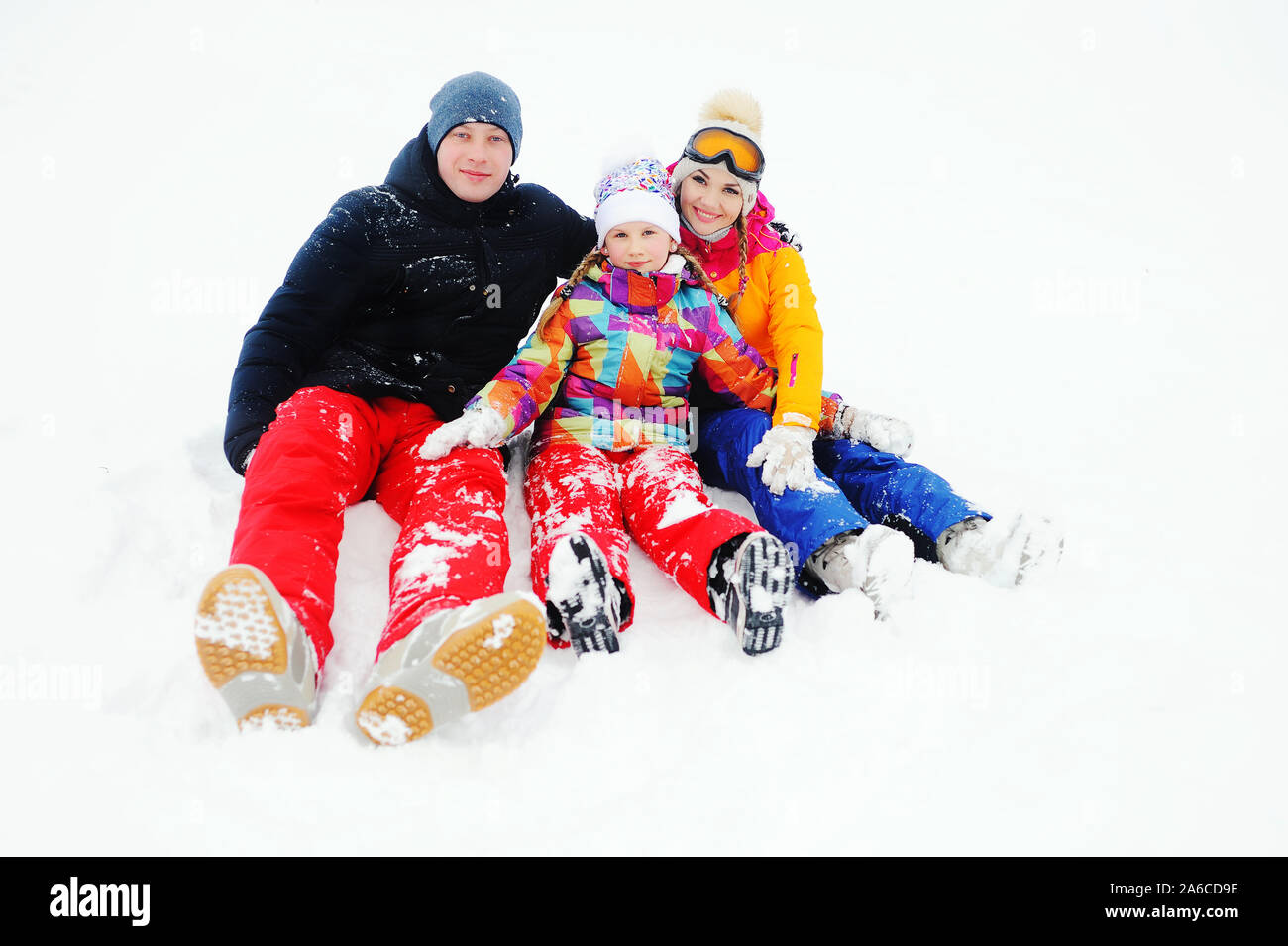 Eine junge Familie - Mutter, Vater und Tochter in hellen Skianzuege sitzen zusammen im Schnee Lachen und Lächeln Stockfoto